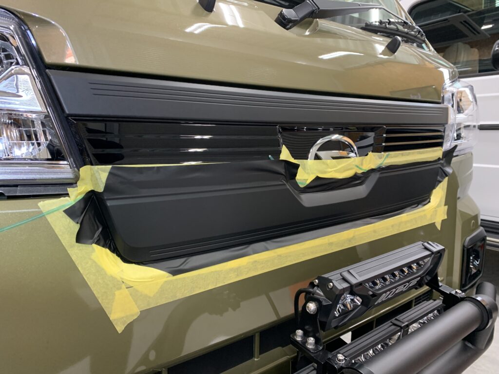 フロントグリルにマットブラックのカーラッピングを施工している途中工程。位置決めとシワ処理を行いながら丁寧に貼り込んでいる様子。