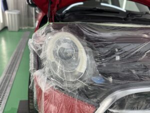 赤いMINIクーパーSのヘッドライトに透明プロテクションフィルムを施工している様子。ボンネット周辺を養生し、フィルム施工中の状態。