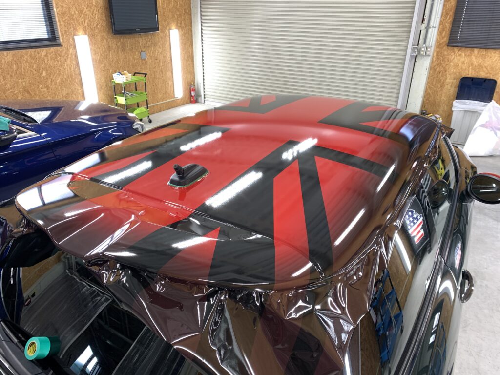 MINI F56 JCWのルーフラッピング施工中の写真。光沢感のあるレッドベースにブラックのユニオンジャックデザインを配置し、立体感のあるルーフ形状に合わせて丁寧に施工している。ルーフスポイラーまで一体感のあるデザインが特徴。