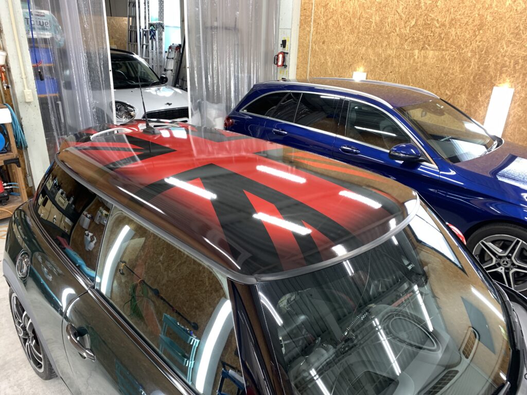 斜め前方から撮影したMINI F56 JCWのルーフラッピング完成写真。CABANAデザインをモチーフにしたレッド×ブラックのユニオンジャック柄をルーフ全面へ施工し、MINIらしいスポーティーで個性的な印象に仕上げている。フロント側から見ることで、ルーフ形状に沿って自然につながるデザインラインや光沢感のある仕上がりが際立っている。